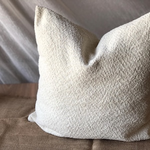 May include: A square throw pillow featuring a textured, off-white boucl&eacute; fabric cover. The material has a distinct nubby, looped surface that creates a tactile appearance. The pillow is plump and maintains a structured shape with defined corners.