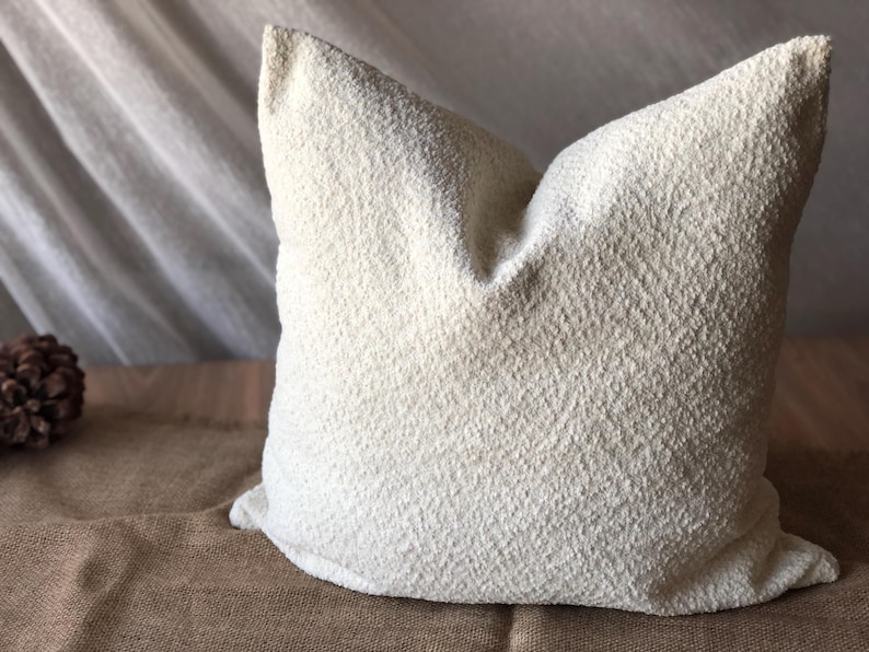 May include: A square throw pillow featuring a textured, off-white boucl&eacute; fabric cover. The material has a distinct nubby, looped surface that creates a tactile appearance. The pillow is plump and maintains a structured shape with defined corners.