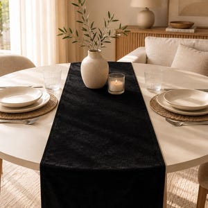 Peut inclure: Un long chemin de table rectangulaire en velours noir centré sur une table à manger ronde blanche. Le chemin de table présente une surface en tissu doux et texturé, placé sous un vase en céramique beige avec des branches d'olivier et une bougie.