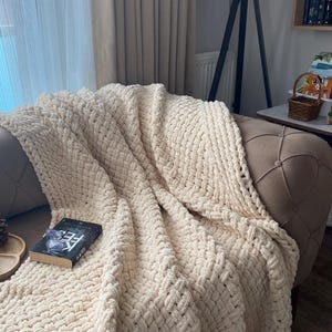 May include: A cream-colored, chunky knit blanket draped over a tufted sofa. The blanket has a woven texture and is folded to reveal its thickness. A book with the title "KARS" is resting on the blanket.