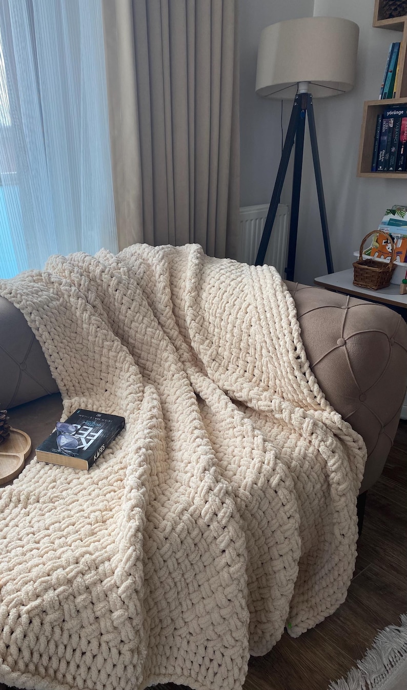 May include: A cream-colored, chunky knit blanket draped over a tufted sofa. The blanket has a woven texture and is folded to reveal its thickness. A book with the title "KARS" is resting on the blanket.