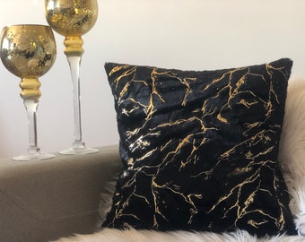May include: Black and gold decorative throw pillow with a marble pattern. The pillow is on a white faux fur surface.