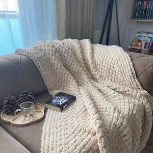 May include: A cream-colored, chunky knit blanket draped over a brown sofa. A book, a wooden tray with a glass mug, and pinecones are on the sofa. The blanket has a woven pattern and a soft, inviting texture.