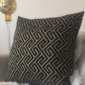 May include: Black decorative throw pillow with a gold geometric pattern. The pillow is square-shaped and rests on a gray surface with a white, fluffy texture. A gold candle holder is visible in the background.