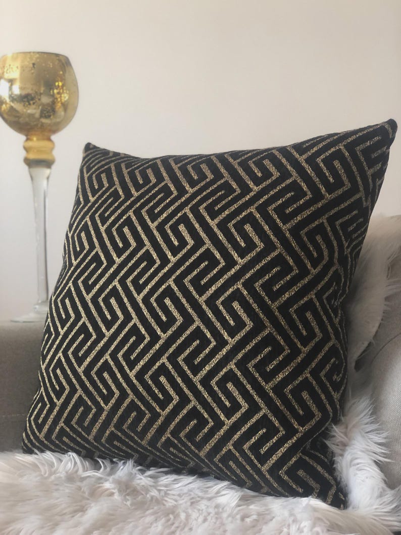 May include: Black decorative throw pillow with a gold geometric pattern. The pillow is square-shaped and rests on a gray surface with a white, fluffy texture. A gold candle holder is visible in the background.
