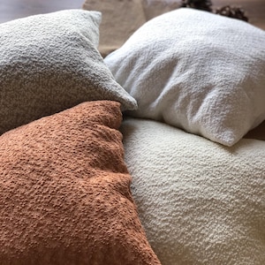 May include: A collection of four square throw pillows featuring a textured, nubby fabric surface. The pillows are presented in a variety of neutral and warm earth tones, including shades of cream, off-white, light gray, and a deep terracotta orange.