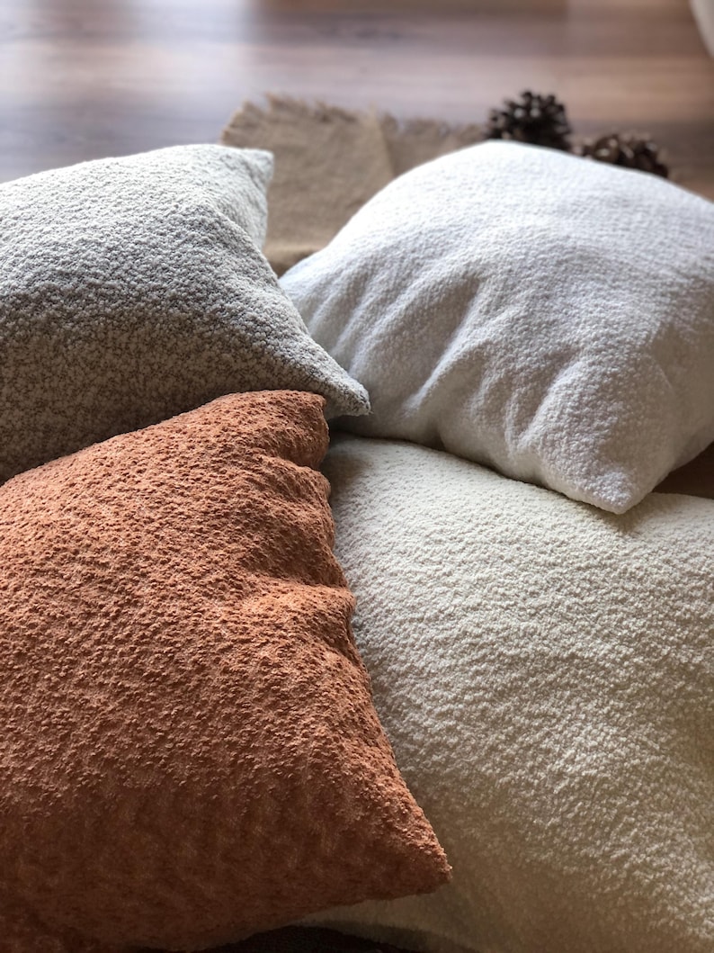 May include: A collection of four square throw pillows featuring a textured, nubby fabric surface. The pillows are presented in a variety of neutral and warm earth tones, including shades of cream, off-white, light gray, and a deep terracotta orange.