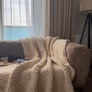 May include: A cream-colored, chunky knit blanket draped over a tufted sofa. The blanket's thick weave is visible, creating a cozy texture. The sofa is positioned near a window with sheer curtains, and a floor lamp is in the background.