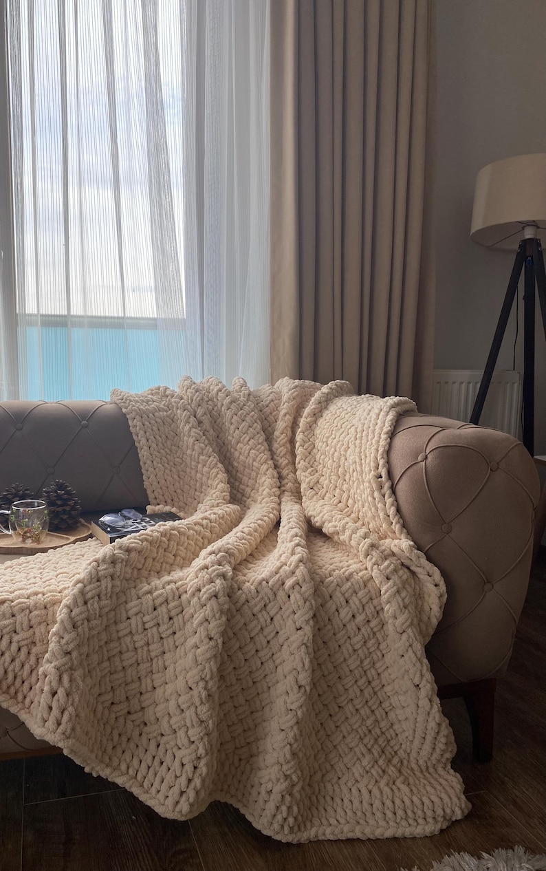 May include: A cream-colored, chunky knit blanket draped over a tufted sofa. The blanket's thick weave is visible, creating a cozy texture. The sofa is positioned near a window with sheer curtains, and a floor lamp is in the background.