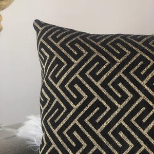 May include: Black decorative pillow with a gold geometric pattern. The pillow has a square shape and a textured surface. The gold pattern is a repeating Greek key design.