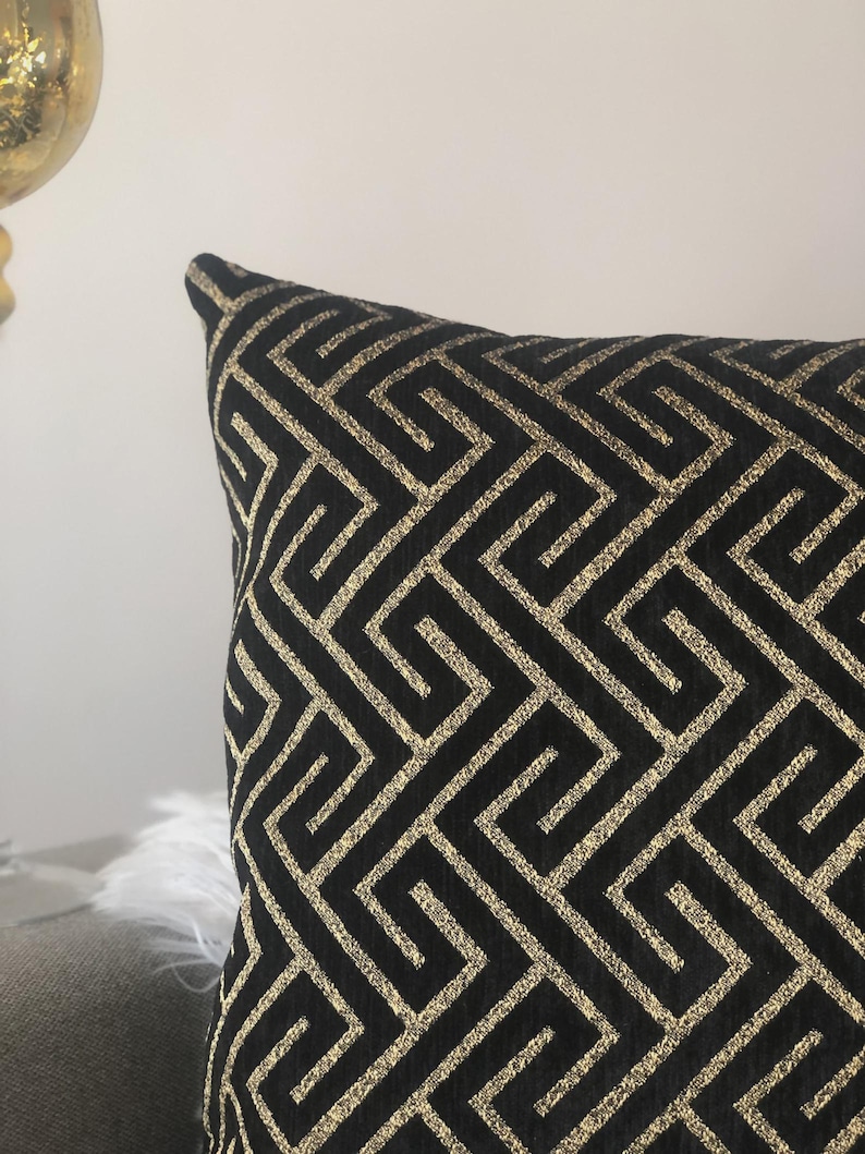 May include: Black decorative pillow with a gold geometric pattern. The pillow has a square shape and a textured surface. The gold pattern is a repeating Greek key design.