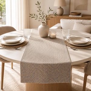 May include: A dining table set with a woven table runner, white plates, and glasses. A vase with greenery and a lit candle are on the table. The round table has a light wood base. The table runner is approximately 150 cm long.