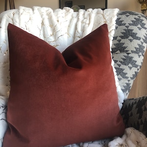 May include: A rust-colored velvet pillow on a white faux fur throw blanket. The pillow is square and has a simple design.