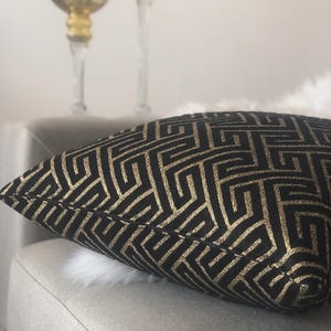 May include: A decorative pillow with a black background and a gold geometric pattern. The pillow is placed on a light gray chair. Two gold-colored candle holders are in the background.