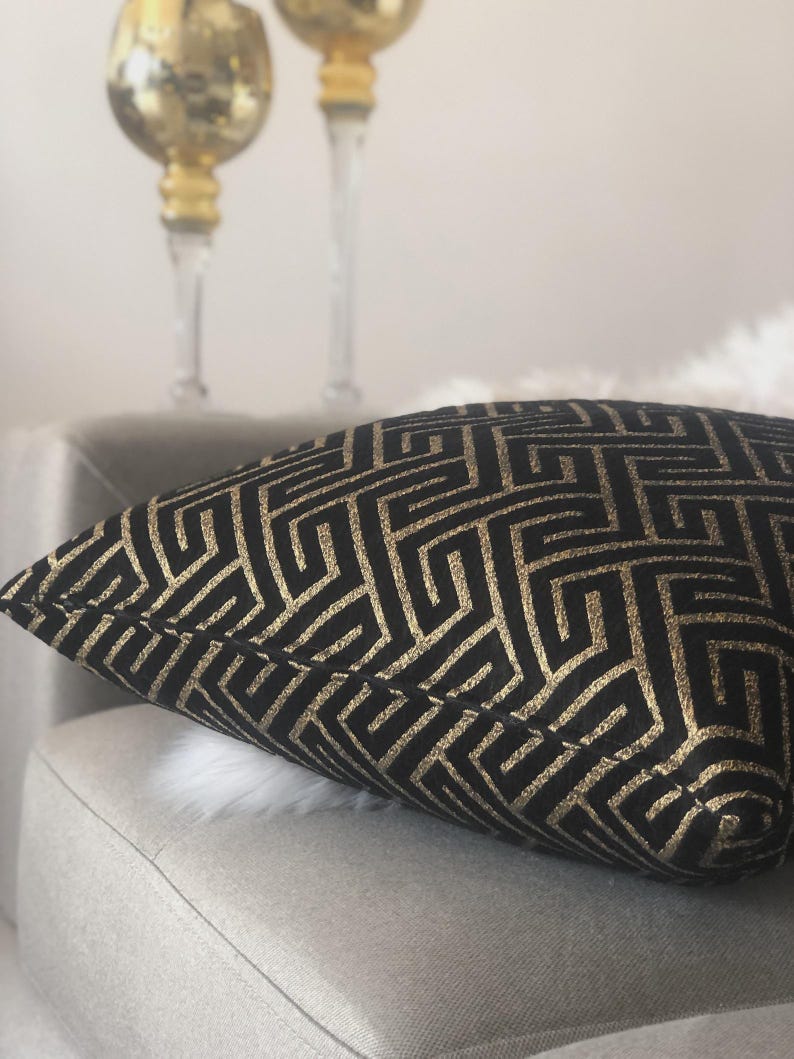 May include: A decorative pillow with a black background and a gold geometric pattern. The pillow is placed on a light gray chair. Two gold-colored candle holders are in the background.