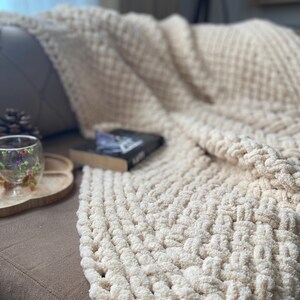 May include: A cream-colored, chunky knit blanket draped over a brown sofa. The blanket has a textured, woven pattern. A book and a decorative glass with floral accents sit on a wooden tray nearby. A cozy, inviting scene.