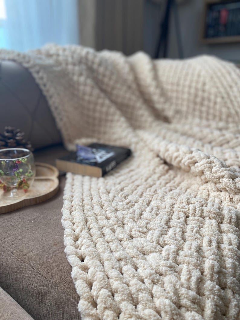 May include: A cream-colored, chunky knit blanket draped over a brown sofa. The blanket has a textured, woven pattern. A book and a decorative glass with floral accents sit on a wooden tray nearby. A cozy, inviting scene.