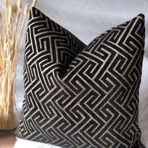 May include: A decorative throw pillow with a black background and a gold geometric pattern. The pillow is square-shaped and rests on a white, fluffy surface. The pillow's design features a repeating maze-like pattern.