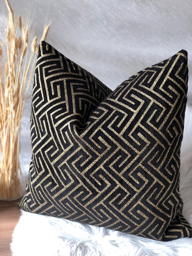 May include: A decorative throw pillow with a black background and a gold geometric pattern. The pillow is square-shaped and rests on a white, fluffy surface. The pillow's design features a repeating maze-like pattern.