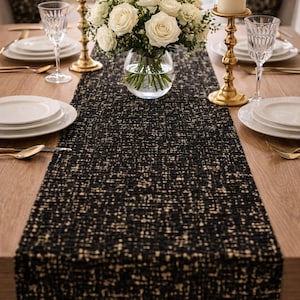 Może przedstawiać: Nakrycie stołu z czarno-beżowym, teksturowanym bieżnikiem. Na stole białe talerze, złote sztućce i kryształowe kieliszki. Bukiet białych róż umieszczony między dwoma zapalonymi świecami w złotych świecznikach.