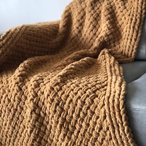May include: A soft, chunky knit throw blanket in a warm mustard yellow color. The blanket is made with a thick yarn and has a woven texture.