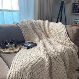 May include: A cream-colored, chunky knit blanket draped over a sofa. The blanket has a thick, woven texture and is folded to create soft, flowing lines. A book with the title "KAFES" is on top of the blanket. A wooden tray with a cup and pinecones is on the sofa.