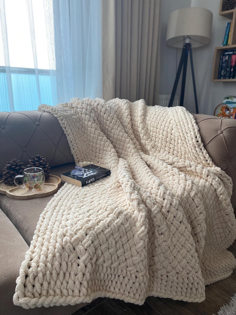 May include: A cream-colored, chunky knit blanket draped over a sofa. The blanket has a thick, woven texture and is folded to create soft, flowing lines. A book with the title "KAFES" is on top of the blanket. A wooden tray with a cup and pinecones is on the sofa.