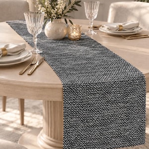 Peut inclure: Un chemin de table rectangulaire présentant un motif tissé texturé en fils noirs et blancs. Le tissu a une apparence dense et mouchetée créée par l'entrelacement de fibres sombres et claires, s'étendant au centre d'une table ronde en bois.