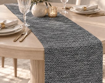 May include: A rectangular table runner featuring a textured, woven pattern in black and white threads. The fabric has a dense, speckled appearance created by the interlacing of dark and light fibers, extending across the center of a round wooden table.