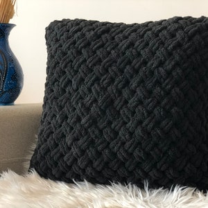 Könnte beinhalten: Schwarzes gestricktes Kissen mit einem strukturierten gewebten Muster. Das Kissen liegt auf einer weißen Kunstfell-Oberfläche.