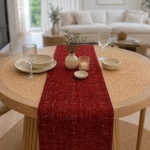 May include: A deep red rectangular table runner with a textured, woven appearance featuring subtle white flecks. The runner is centred on a round wooden table with a woven rattan top, accompanied by two clear ribbed glasses and ceramic bowls.