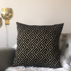 May include: Black and gold geometric patterned throw pillow with a subtle sheen. The pillow is on a white faux fur throw and a gray couch.