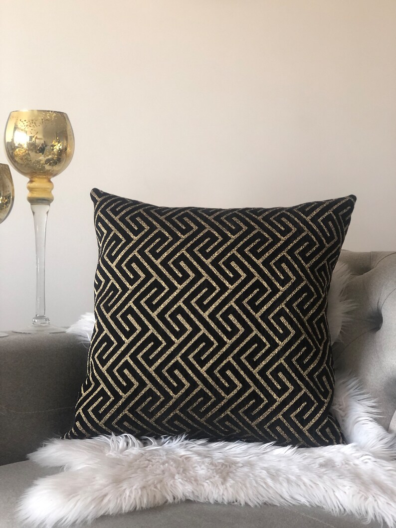 May include: Black and gold geometric patterned throw pillow with a subtle sheen. The pillow is on a white faux fur throw and a gray couch.