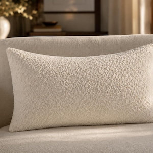 May include: A rectangular throw pillow featuring a textured, cream-colored boucl&eacute; fabric. The surface of the pillow displays a dense, looped weave that creates a consistent, tactile appearance across the entire front face of the cushion.