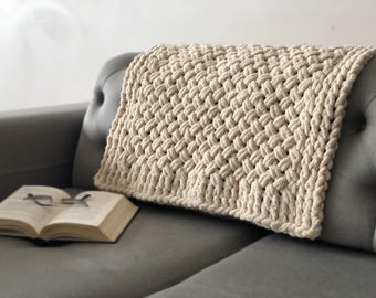 May include: A cream-colored knitted throw blanket with a woven texture is draped over a gray couch. The blanket is made of thick yarn and has a soft, cozy feel.