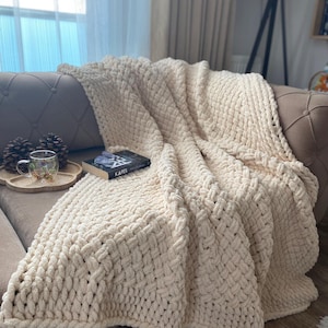 May include: A cream-colored, chunky knit blanket draped over a sofa. The blanket has a thick, textured weave and is folded to create a cozy, inviting look. A book with the title "KAFES" is on top of the blanket.