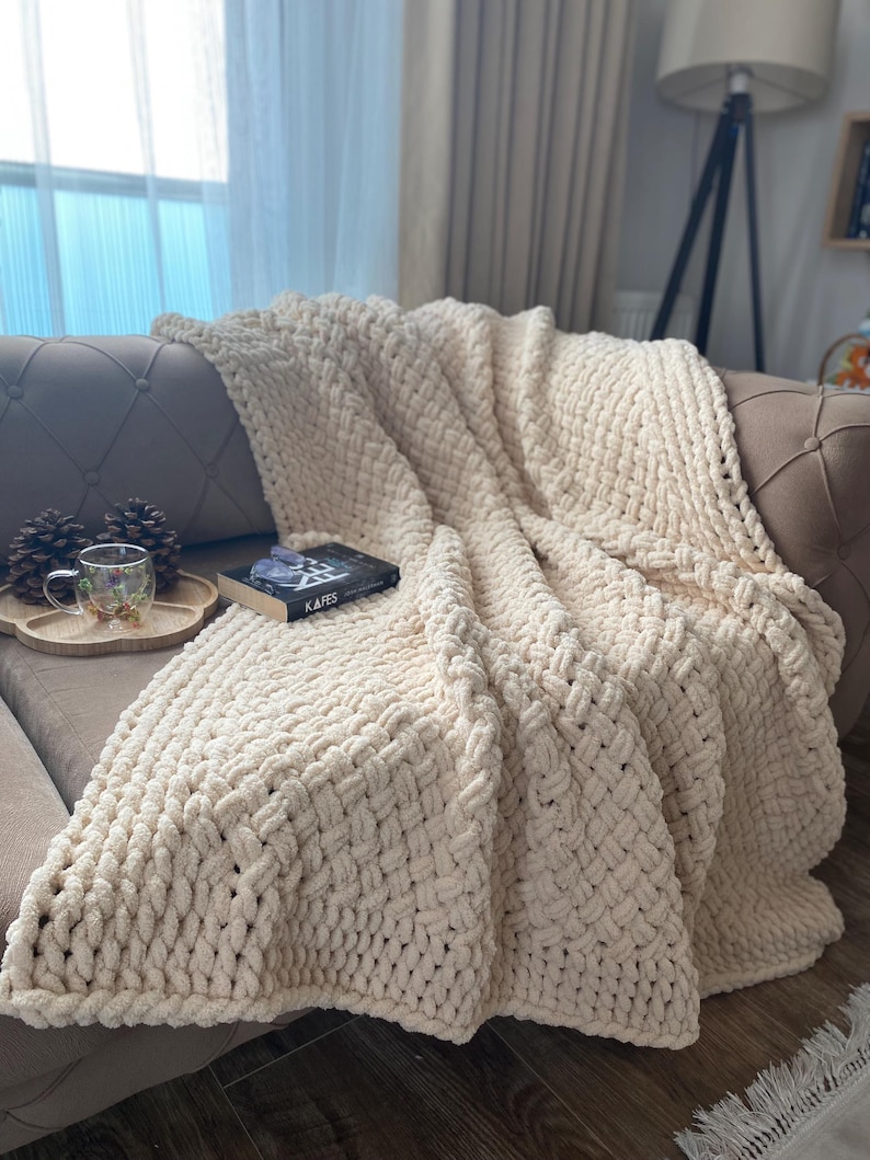 May include: A cream-colored, chunky knit blanket draped over a sofa. The blanket has a thick, textured weave and is folded to create a cozy, inviting look. A book with the title "KAFES" is on top of the blanket.