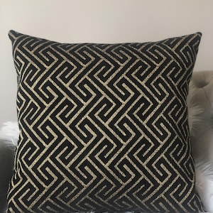 May include: Square black throw pillow with a gold geometric pattern. The pillow has a repeating Greek key design in a metallic gold color. The pillow is on a white furry surface.