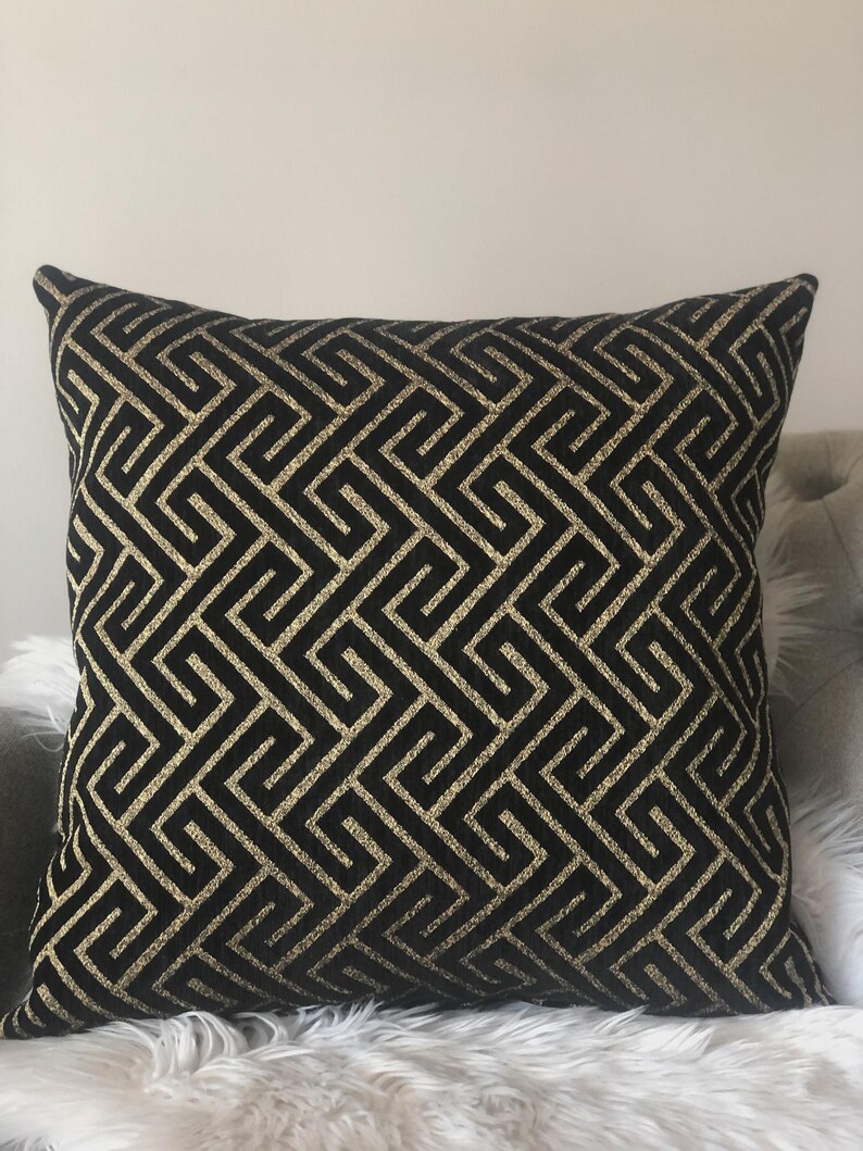 May include: Square black throw pillow with a gold geometric pattern. The pillow has a repeating Greek key design in a metallic gold color. The pillow is on a white furry surface.