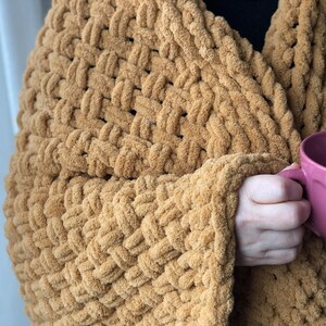 May include: A close-up of a chunky knit blanket in a warm brown color. The blanket is draped over a person's arm, and they are holding a pink mug.