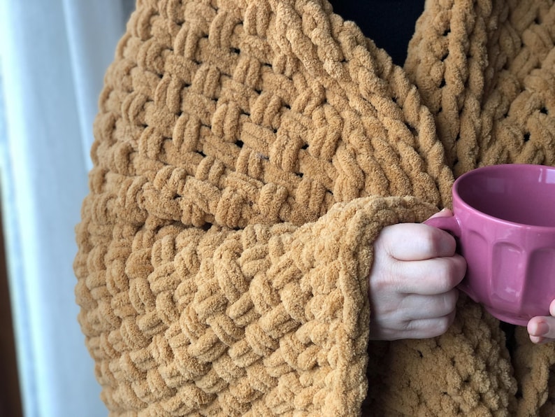 May include: A close-up of a chunky knit blanket in a warm brown color. The blanket is draped over a person's arm, and they are holding a pink mug.