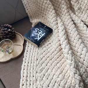 May include: A cream-colored, chunky knit blanket with a woven pattern is draped over a sofa. A book with the title "KAFES" is resting on the blanket. A wooden tray with a pinecone and a glass with flowers is on the sofa.