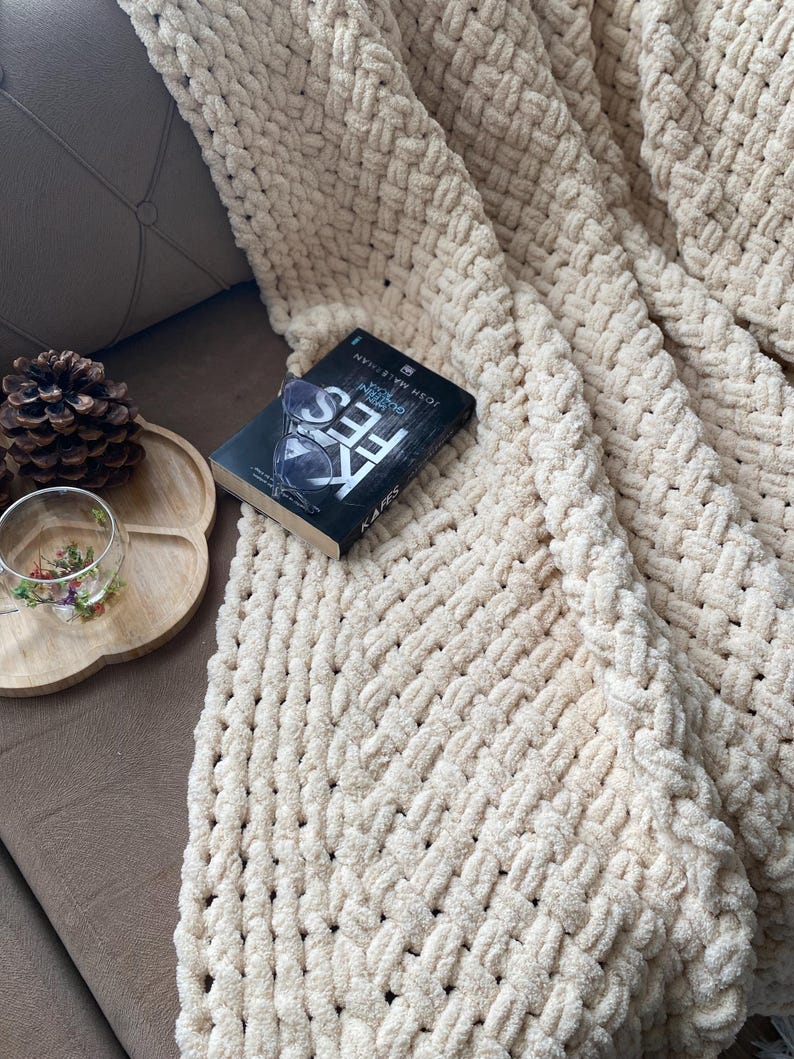 May include: A cream-colored, chunky knit blanket with a woven pattern is draped over a sofa. A book with the title "KAFES" is resting on the blanket. A wooden tray with a pinecone and a glass with flowers is on the sofa.