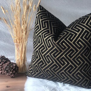 May include: Black and gold geometric patterned throw pillow. The pillow features a repeating maze-like design in gold against a black background. The pillow is placed on a white surface with decorative wheat stalks in a clear vase.