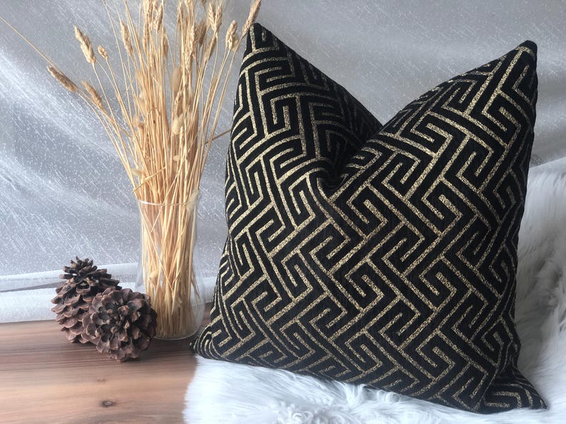 May include: Black and gold geometric patterned throw pillow. The pillow features a repeating maze-like design in gold against a black background. The pillow is placed on a white surface with decorative wheat stalks in a clear vase.