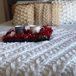 May include: Two mugs with holiday designs sit on a tray with red tinsel and ornaments. The tray is on a white fluffy blanket on a bed with pillows.