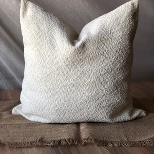 May include: A square throw pillow featuring a textured, off-white boucl&eacute; fabric surface. The pillow has a soft, nubby appearance with a consistent cream-colored finish across the entire front face, showcasing a dense, looped textile construction.