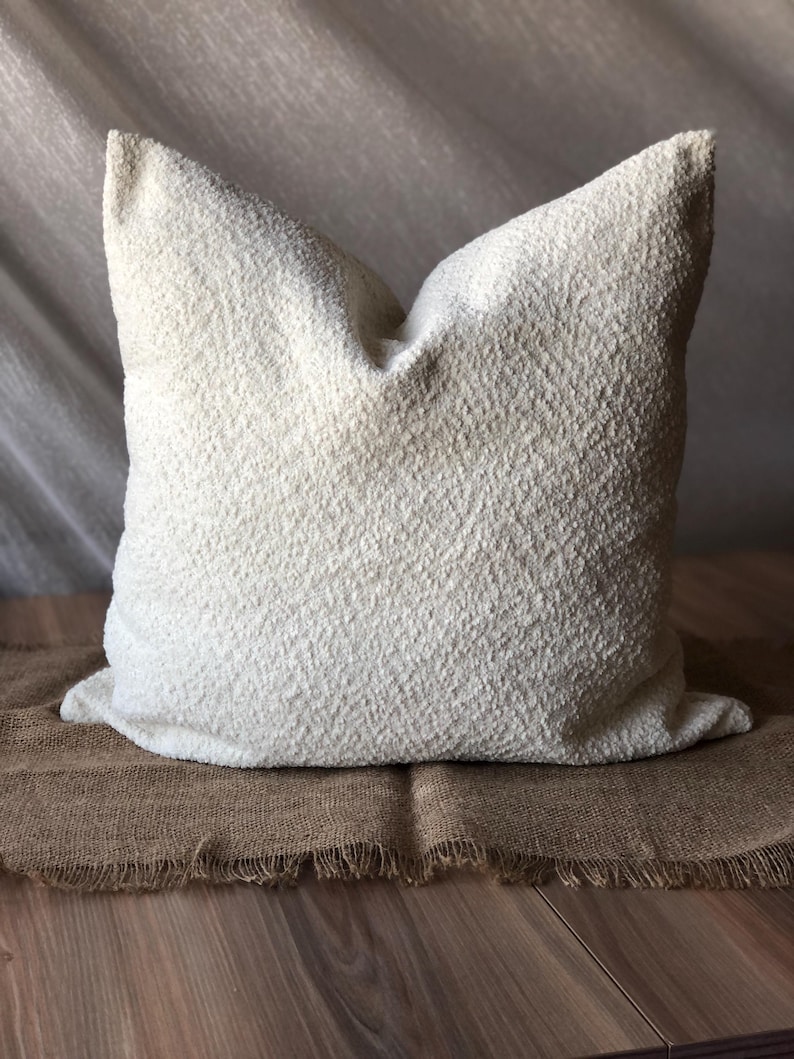 May include: A square throw pillow featuring a textured, off-white boucl&eacute; fabric surface. The pillow has a soft, nubby appearance with a consistent cream-colored finish across the entire front face, showcasing a dense, looped textile construction.