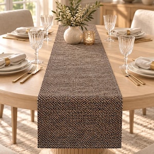 May include: A rectangular table runner featuring a dark brown and black woven texture with a subtle speckled pattern. The runner is centred on a round light wood dining table, extending across the surface between place settings and decor.