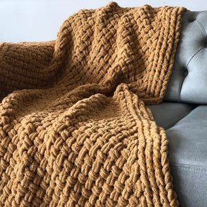 May include: A chunky knit throw blanket in a warm brown color. The blanket is made with a thick yarn and has a textured weave. It is draped over a gray couch.
